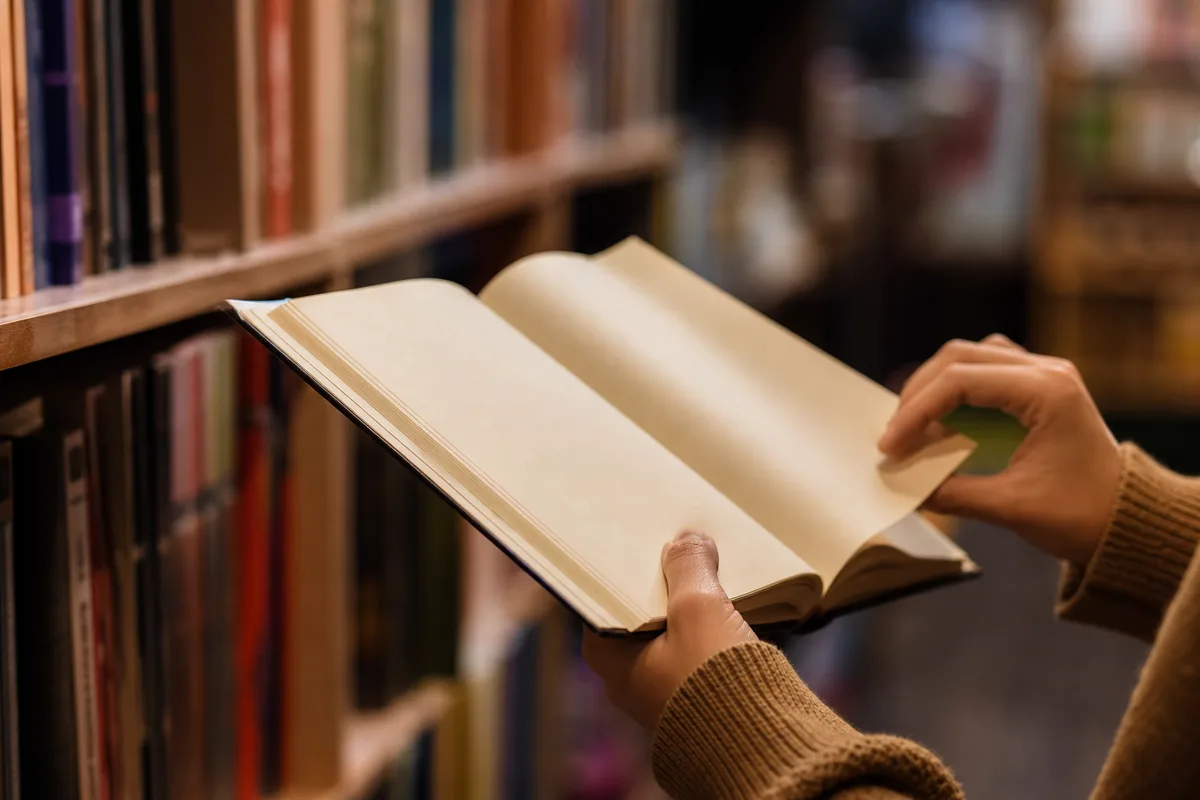Mains feuilletant un roman ouvert dans une librairie, rayonnages de livres flous en arrière-plan
